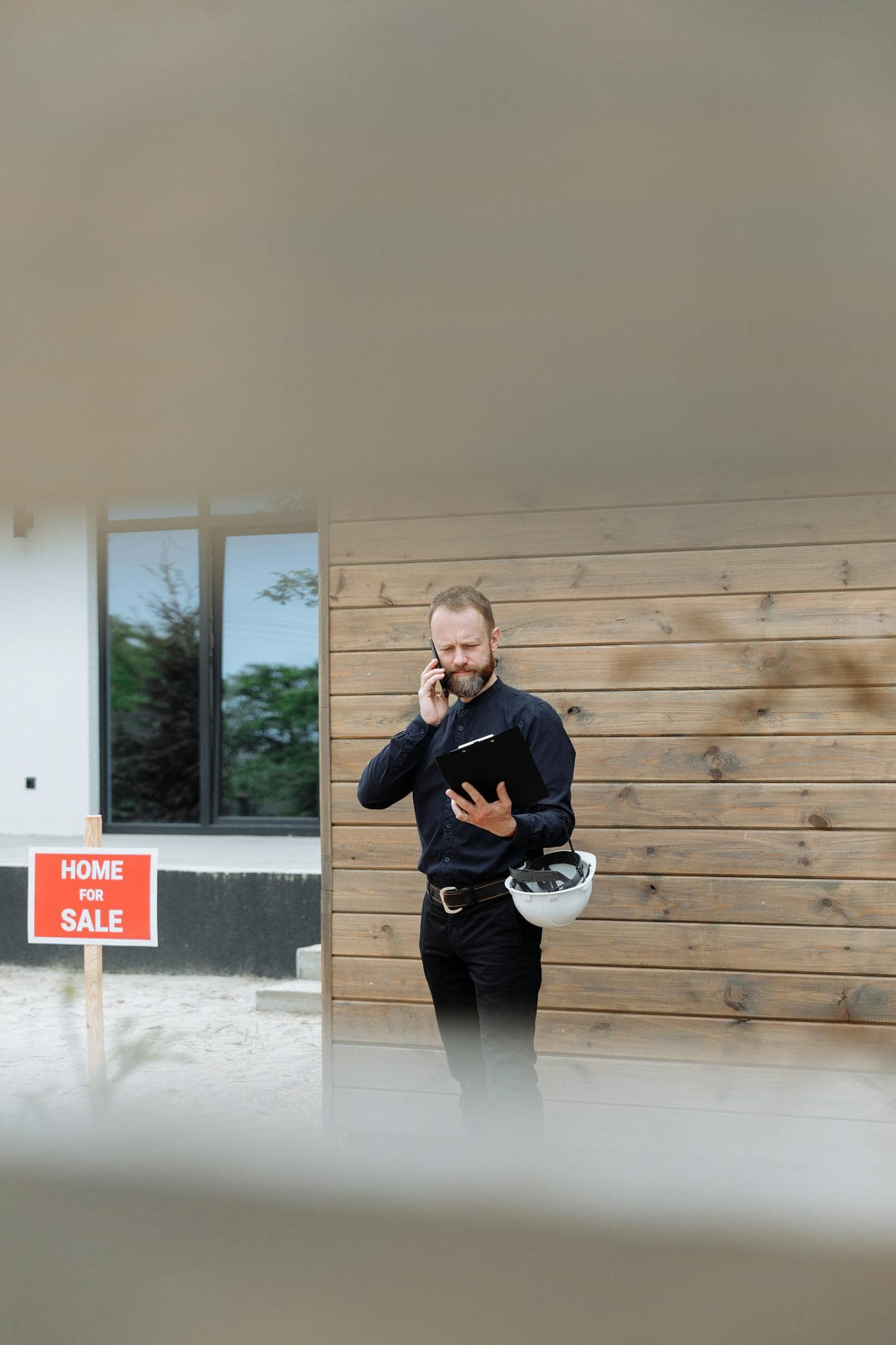 Real estate agent holding phone and clipboard in front of a house with for sale sign.